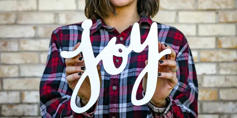 Frau hält das Wort "Joy" aus Holz in der Hand vor einer Backsteinwand, trägt ein kariertes Hemd.
