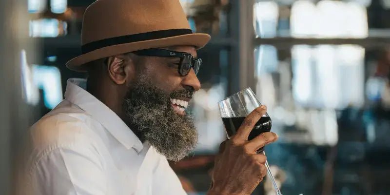 Wein: Genießer mit Hut und Brille lächelt beim Verkosten eines Rotweins in einer stilvollen Bar.
