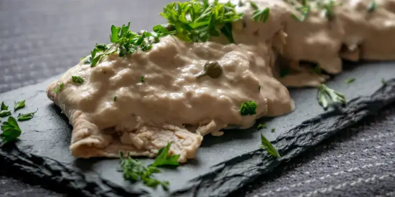 Vitello Tonnato mit Kapern und Thunfischsauce auf schwarzem Teller, garniert mit frischen Kräutern.