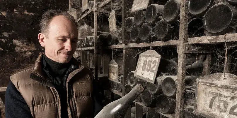 J. Neus mit einer Flasche Wein in einem historischen Weinkeller, umgeben von Fuderfässern und Etiketten.