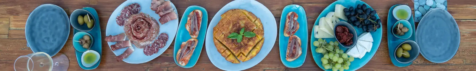 Feinkostübersicht mit Wurst, Käse, Oliven und Früchten auf blauen Tellern präsentiert.