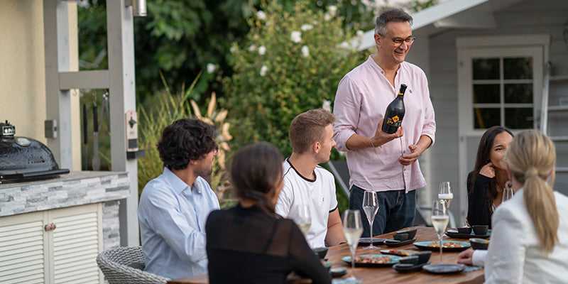 Geselliger Abend im Freien: Gastgeber präsentiert Wein vor Freunden am Tisch, umgeben von Natur.