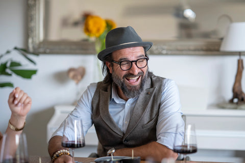 Lachender Mann mit Brille und Hut an einem Tisch, interagiert fröhlich bei einem Essen mit Freunden.
