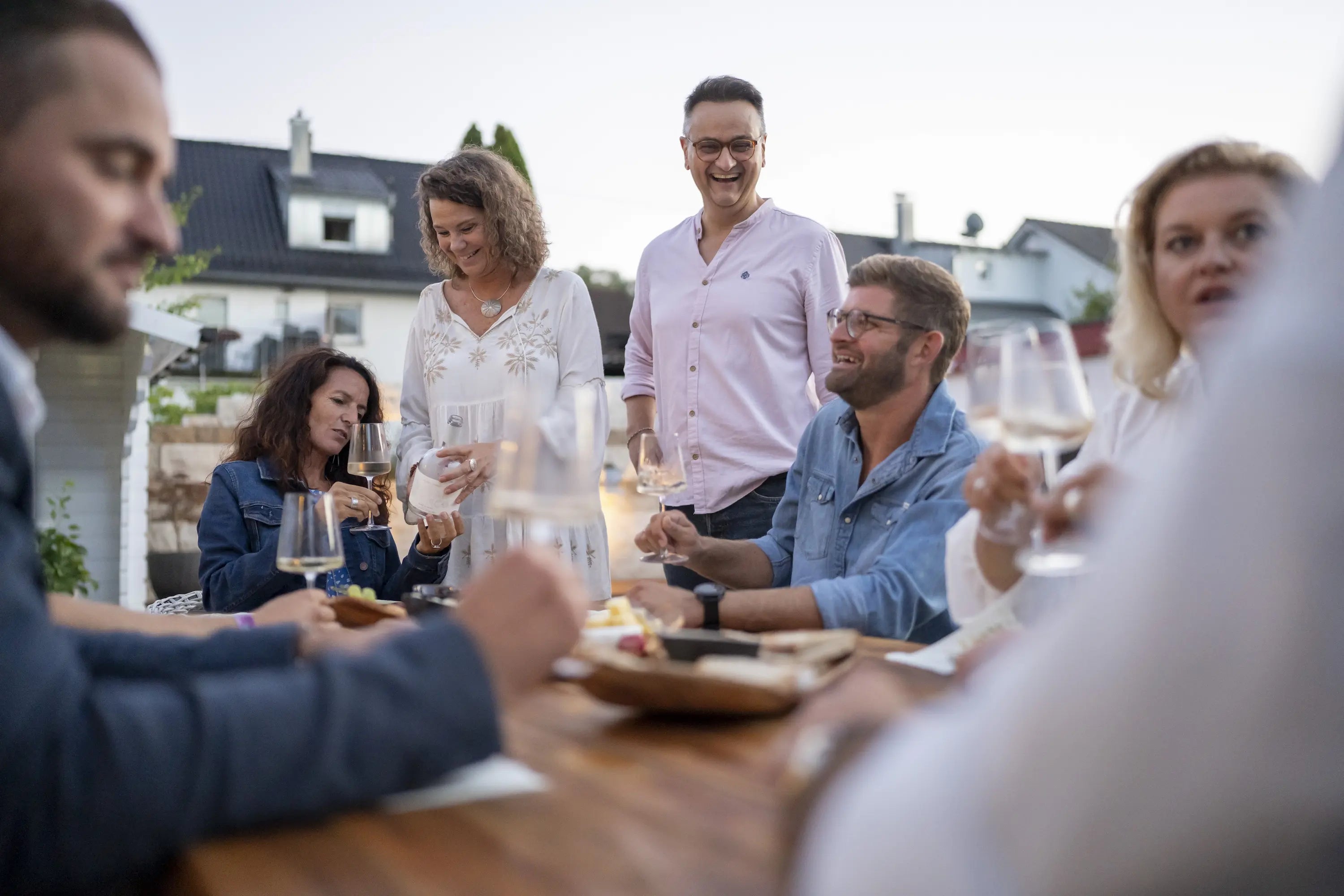 Mehrere lachende Personen in legerer Kleidung sitzen an einem gedeckten Tisch im Garten und prosten sich mit Weingläsern zu. Nadja und Brano stehen neben dem Tisch. 