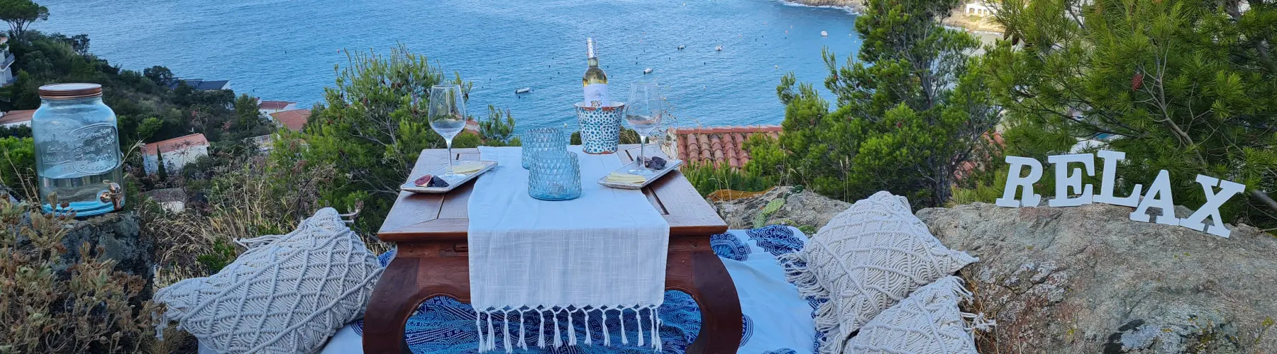 Auf einem Felsen mit Blick aufs Meer steht ein mit Essen und Wein gedeckter Holztisch. Daneben sind Kissen auf dem Boden