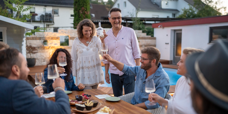 Mehrere lachende Personen in legerer Kleidung sitzen an einem gedeckten Tisch im Garten und prosten sich mit Weingläsern zu. Nadja und Brano stehen neben dem Tisch. Im Hintergrund ist Pool zu sehen.