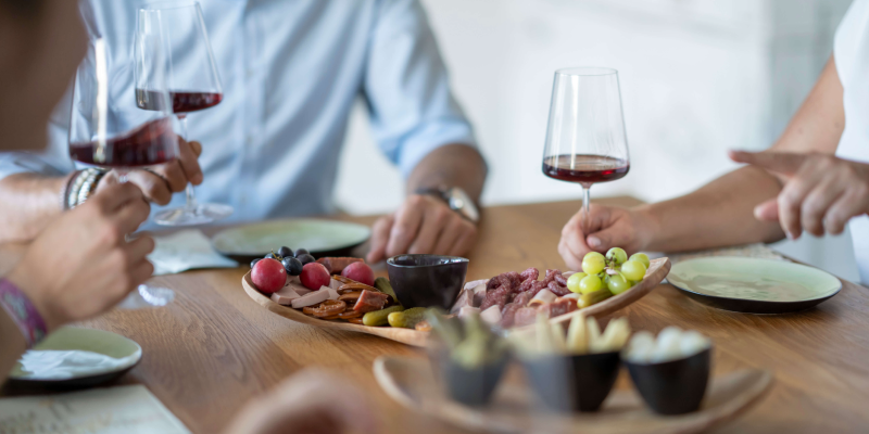 Mehrere Personen sitzen an einem mit Fingerfood gedeckten Esstisch und halten ein Glas mit Rotwein in ihrer Hand