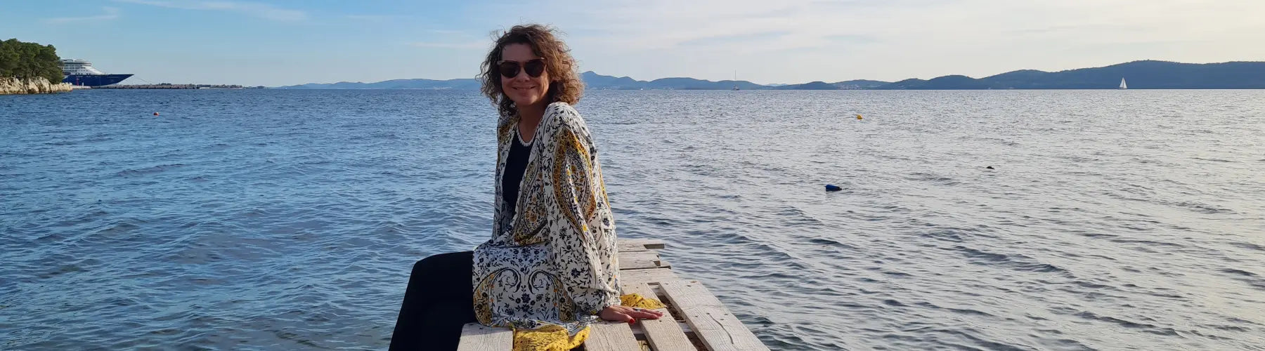 Frau mit Sonnenbrille sitzt am Ufer, Blick auf Wasserlandschaft und Berge im Hintergrund, entspannte Stimmung.