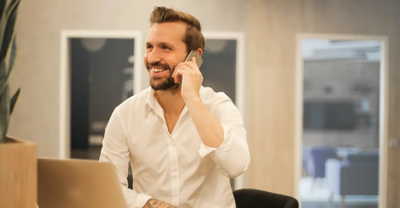 Ein Mann dunkelhaariger Mann mit Vollbart trägt ein weißes Hemd und sitzt in einem Büro, telefoniert und lächelt dabei