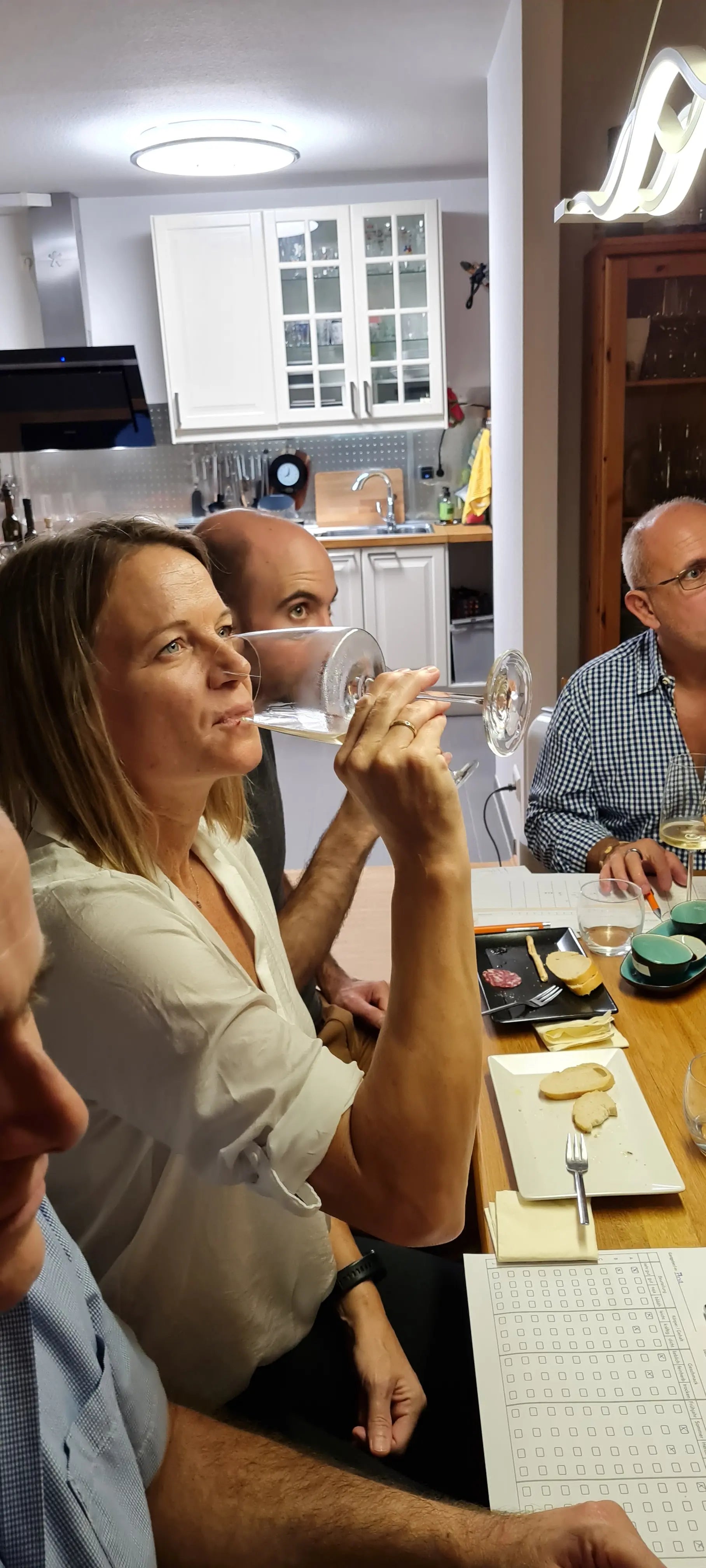 Eine Frau und drei Männer am gedeckten Tisch zum Tasting zusammen. Eine Frau trinkt aus Ihrem Weinglas Tasting. Im Hintergrund sieht man eine weiße Küche