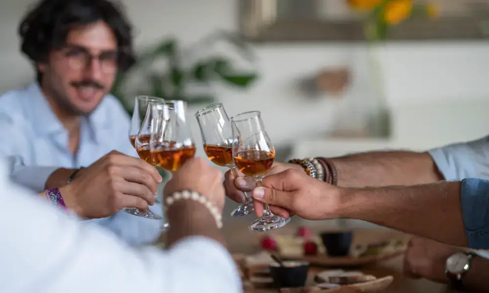 Fünf Personen stoßen mit Whiskygläsern an. In der Bildmitte sind die befüllten Gläser zu sehen, im Hintergrund ist als einziger ein dunkelhaariger Mann mit Brille und Oberlippenbart. 