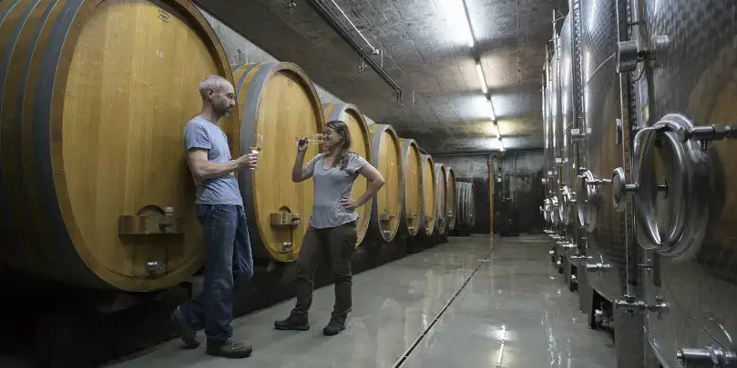 Weingut Meiser: Zwei Personen stehen in einem Weinkeller zwischen großen Holzfässern und diskutieren über Wein.