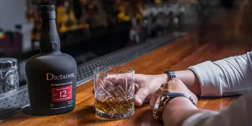 Dictador 12 Jahre Rum in einer Bar, neben einem Glas mit Rum und einer Person, die eine Uhr trägt.