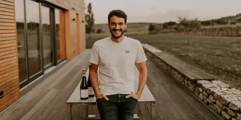 Chardonnay vom Weingut Winter: Stefan Winter steht lächelnd vor dem Weingut mit einer Weinflasche auf der Holzterrasse.