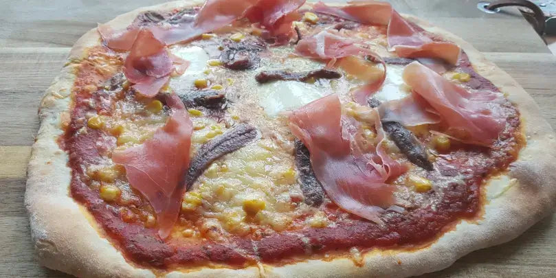Pizza mit Schinken und Sardellenfilets, frisch gebacken auf Holzbrett, appetitlicher Belag und goldener Käse.
