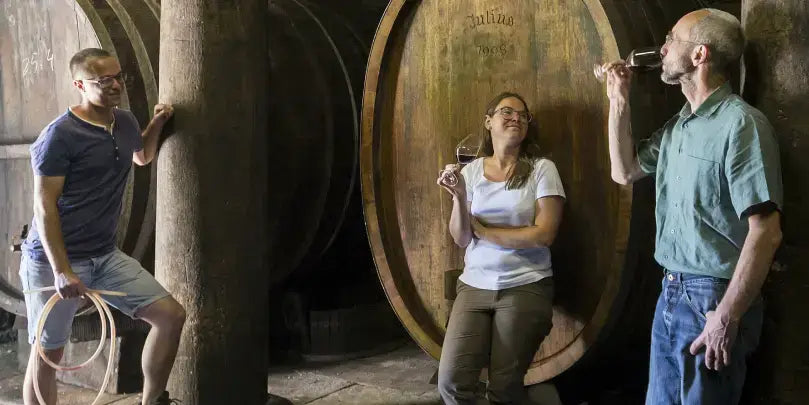 Weingut Meiser: Weinverkostung inmitten alter Holzfässer mit drei Personen, die Wein probieren.