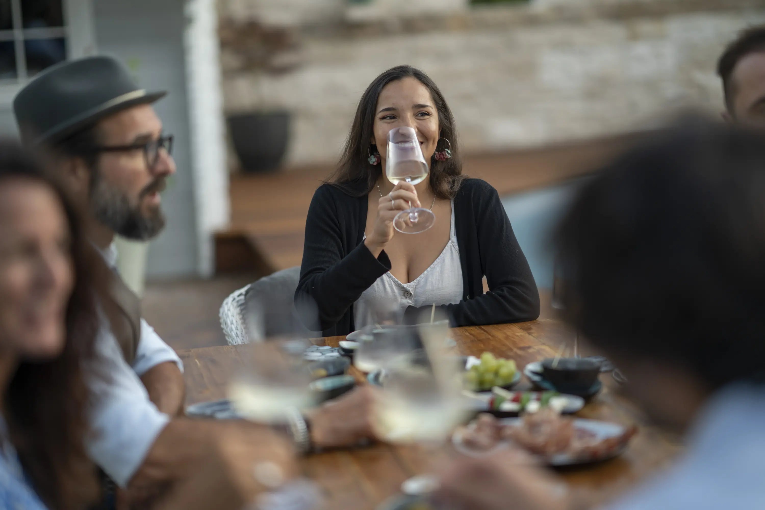 Mehrer Personen sitzen gut gekleidet und fröhlich im Garten an einem Esstisch. Eine junge Frau hält ihr Glas vor das Gesicht und ist fröhlich. Auf dem Esstisch sind auf mehreren Tellern und Schalen Tapas vorhanden. Im Hintergrund ist ein Gartenhaus und ein Gebüsch zu sehen.